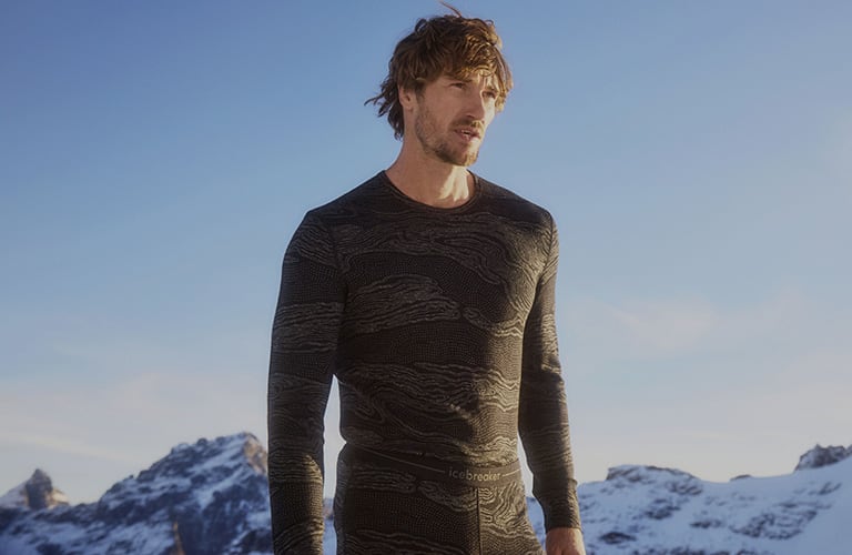 Man standing on snow covered mountain top wearing icebreaker merino base layer thermal top and merino leggings