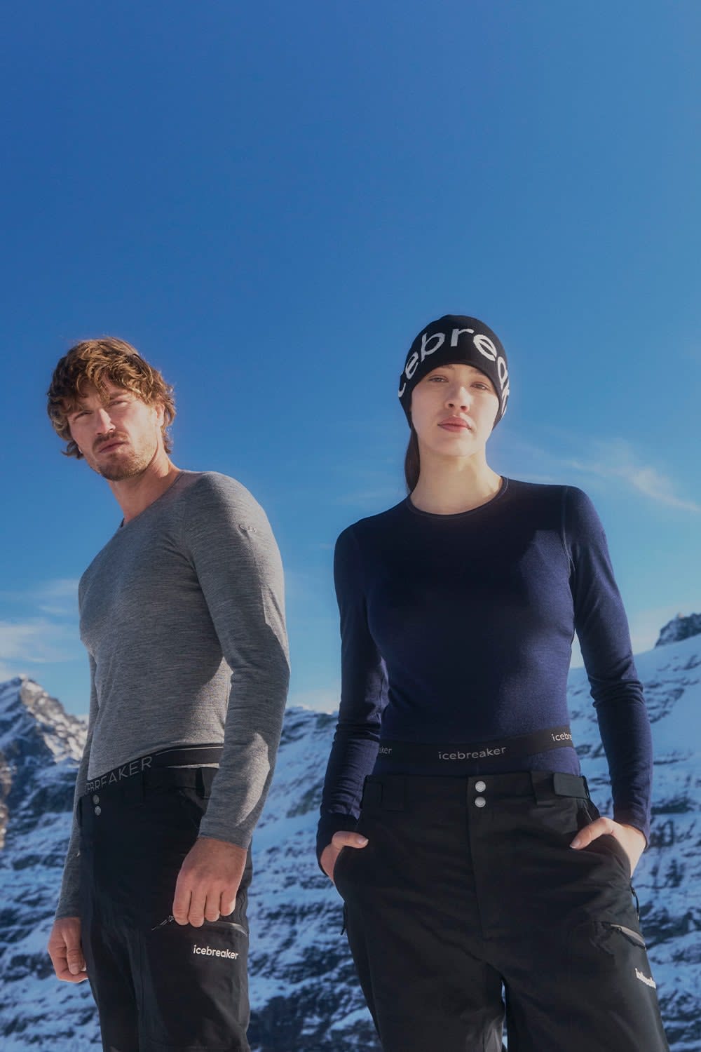 The image features two individuals standing in front of a mountainous landscape with clear blue skies. Both individuals are wearing Icebreaker apparel. The person on the left is wearing a light grey long-sleeved top and black pants with the Icebreaker logo visible on the waistband. The person on the right is dressed in a dark navy long-sleeved top and black pants, also with the Icebreaker logo on the waistband. Behind them, snow-covered peaks rise under the bright sky, suggesting a cold but sunny environment suitable for outdoor activities.