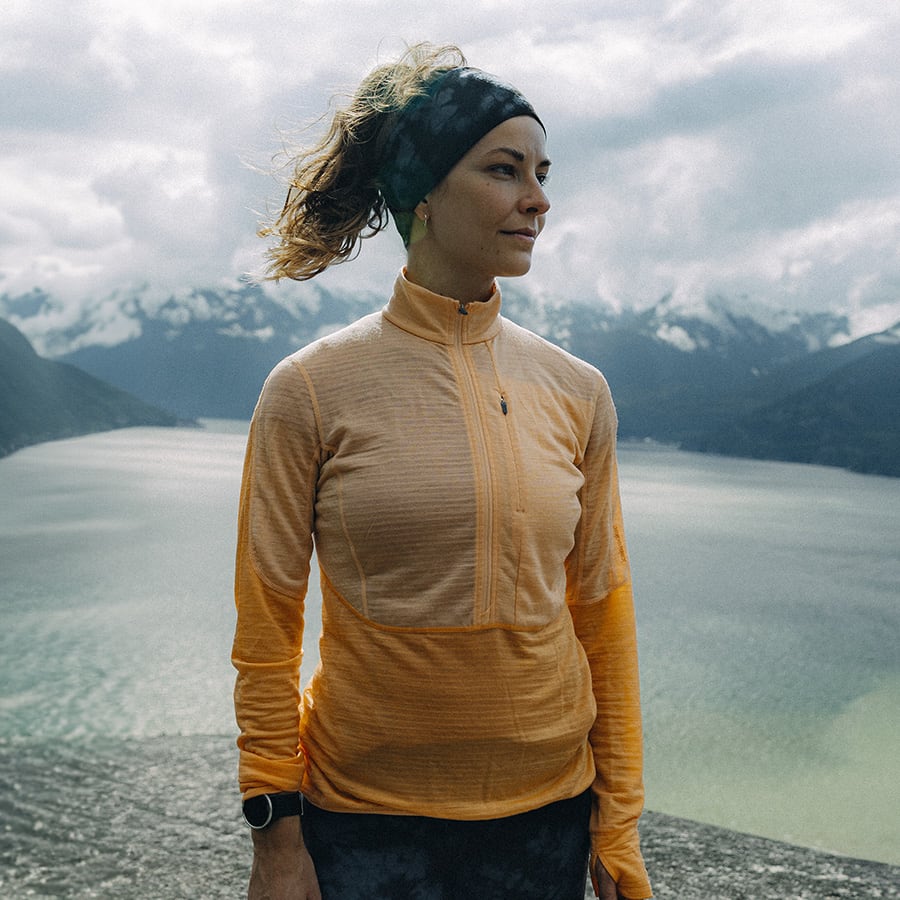 A woman standing on a hill overlooking a lake. She is facing the camera and is wearing an orange icebreaker mid layer. 