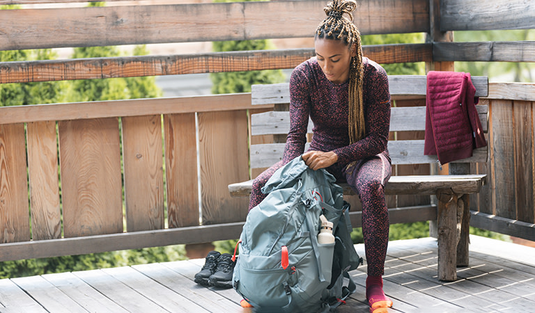 Woman sitting on bench seat packing a hiking backpack wearing icebreaker merino base layer thermal top and leggings