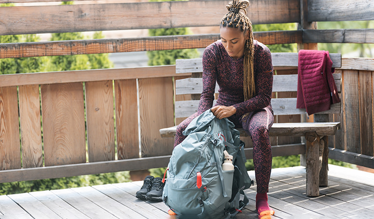 Femme assise sur une banquette emballant un sac à dos de randonnée portant une couche de base thermique en mérinos icebreaker et des leggings